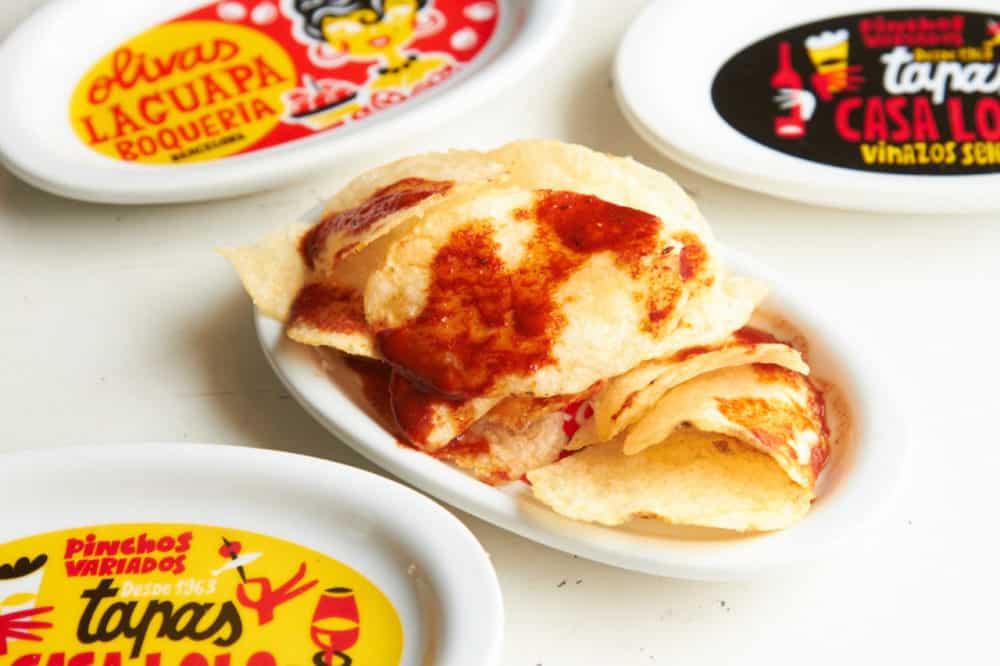 Paprika chips on a small plate surrounded by plates that are red, black and yellow with Spanish phrases written on them.