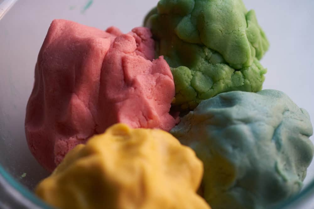 A glass bowl containing four portions of cookie dough in four colors: red, blue, green and yellow.