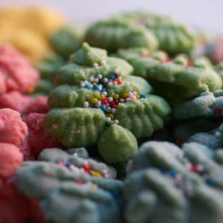 Colorful Christmas cookies o f various shapes and colors, including red wreaths, gold bells, blue ornaments and green Christmas trees. A green Christmas tree-shaped cookie with multicolored non pareils is centered.