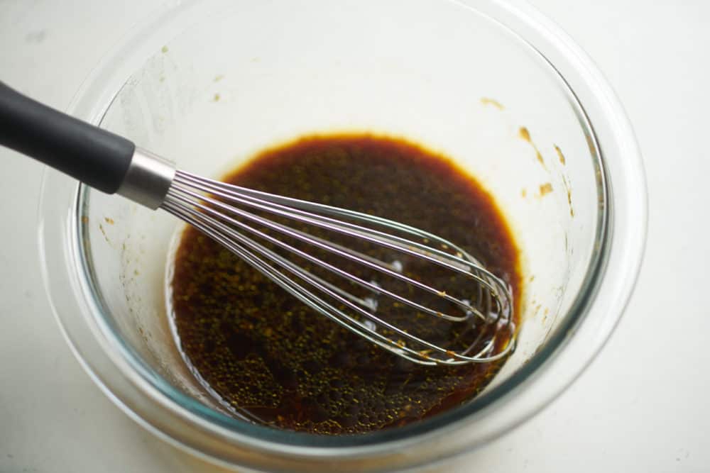 A glass bowl containing soy sauce marinade, shown a whisk on a white surface. 