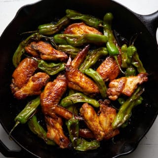 Baked chicken wings with honey, harissa, and shishito peppers in a cast iron skillet displayed on a white surface.