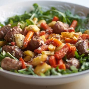 A white bowl with arugula topped with roasted Italian sausages, carrots, and parsnips.