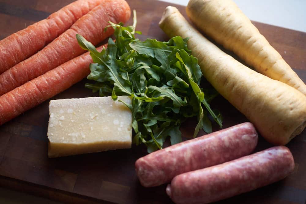 Fresh carrots, parsnips and arugula, alongside Italian sausages and a block of parmesan cheese.