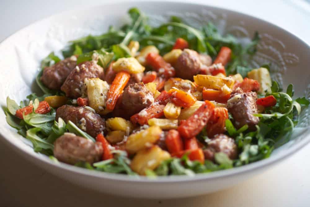 A white bowl with arugula topped with Italian sausages and roasted carrots and parsnips.