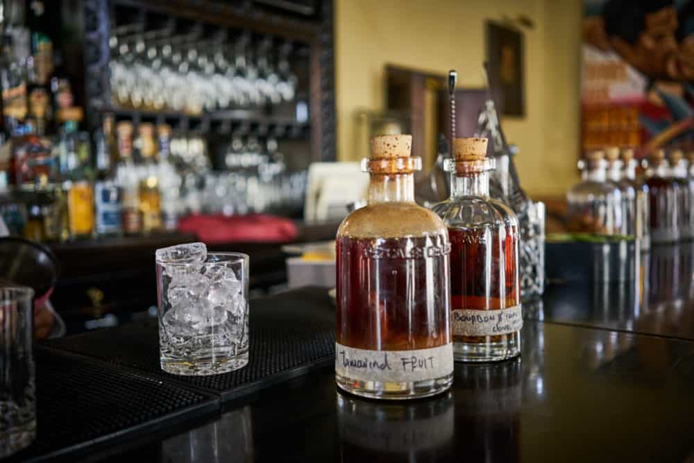 Two glass bottles of infused syrups for cocktails at the Todos Santos Inn, sitting on a wooden bar with liquor bottles in the background. 