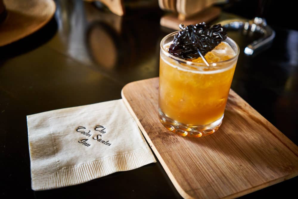 A mezcal cocktail infused with chile sits on a tray on the bar, a cocktail napkin that reads "Todos Santos Inn" sits next to it. Photo by Brian Cummings. 