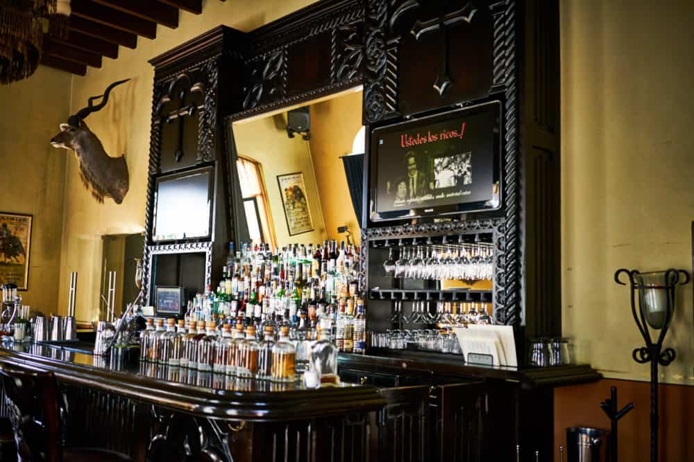 The bar at the Todos Santos Inn features a large wooden bar with mirrors and elaborate woodwork. The room has yellow walls and a deer head, and beams on the ceiling. 