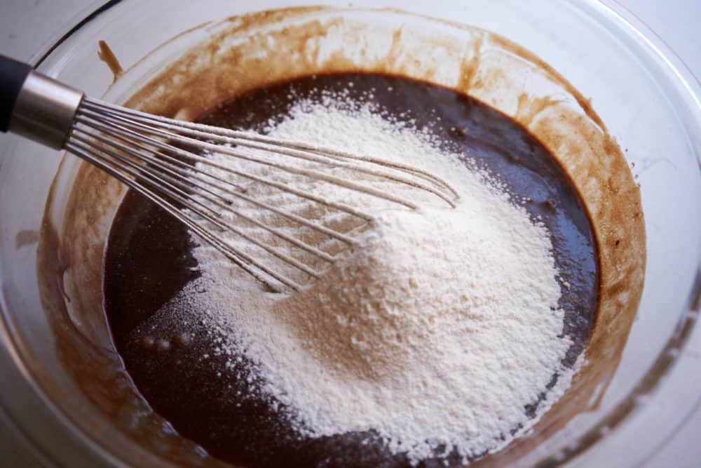 A whisk in a glass bowl containing a chocolate mixture, with sifted flour on top.