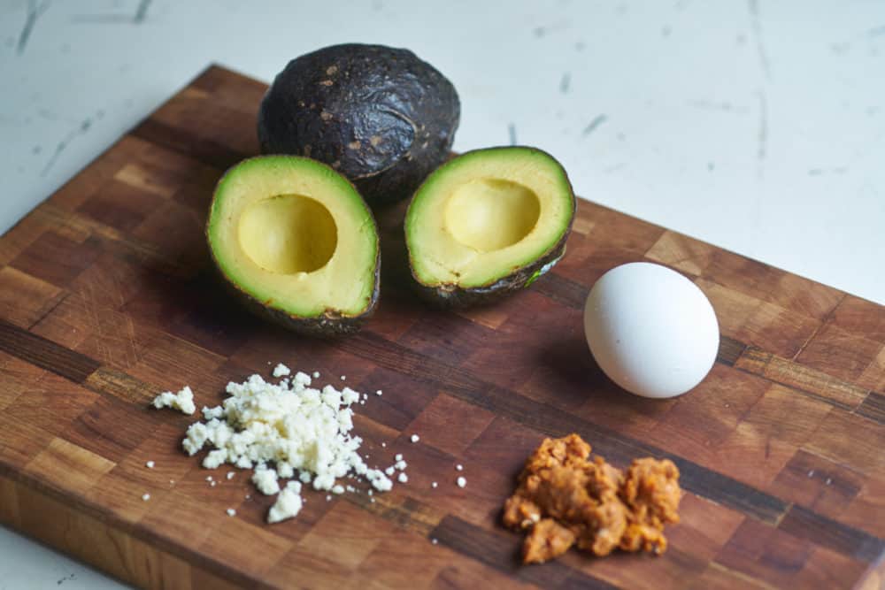 A split avocado, an egg, queso fresco and chorizo displayed on a wooden cutting board.