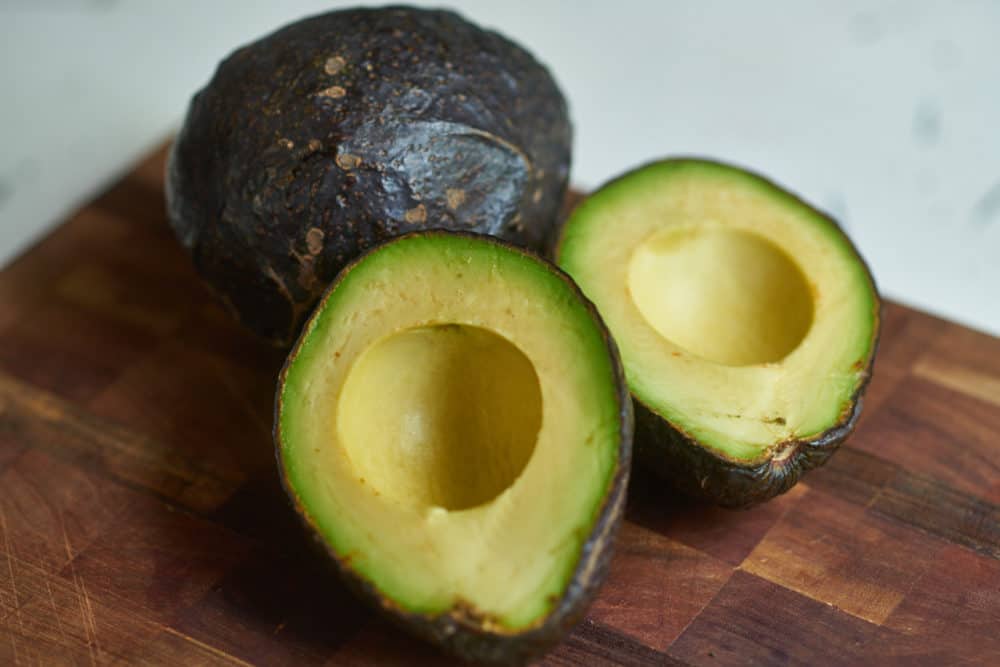 A halved avocado and a whole avocado arranged on a wooden cutting board.