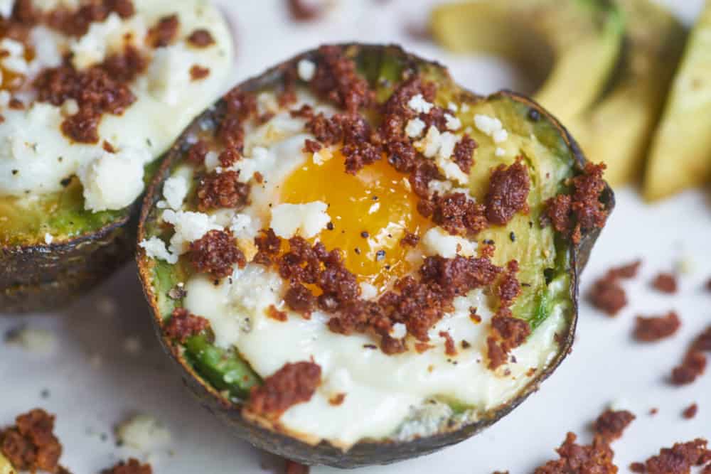 Egg baked in an avocado half topped with chorizo and queso fresco displayed on a white plate, surrounded by bits of chorizo and avocado slices.