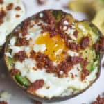 Egg baked in an avocados half topped with chorizo and queso fresco displayed on a white plate, surrounded by bits of chorizo and avocado slices.