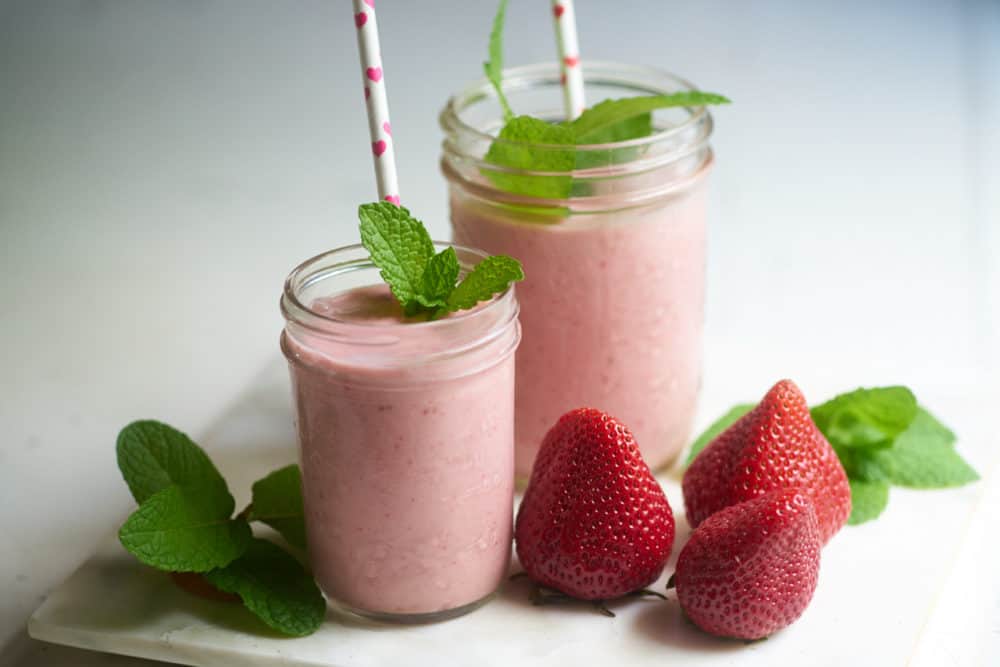 Two dairy-free strawberry shakes in two different sizes of glass ball jars, garnished with mint and paper straws with red hearts. The glasses are sitting on a marble cutting board surrounded by fresh strawberries and sprigs of mint.