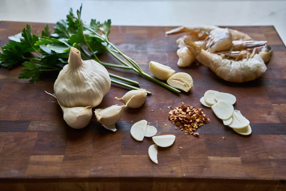 Parsley, a head of garlic, thinly sliced garlic cloves, crushed red pepper, and raw shrimp are displayed on a wooden cutting board.