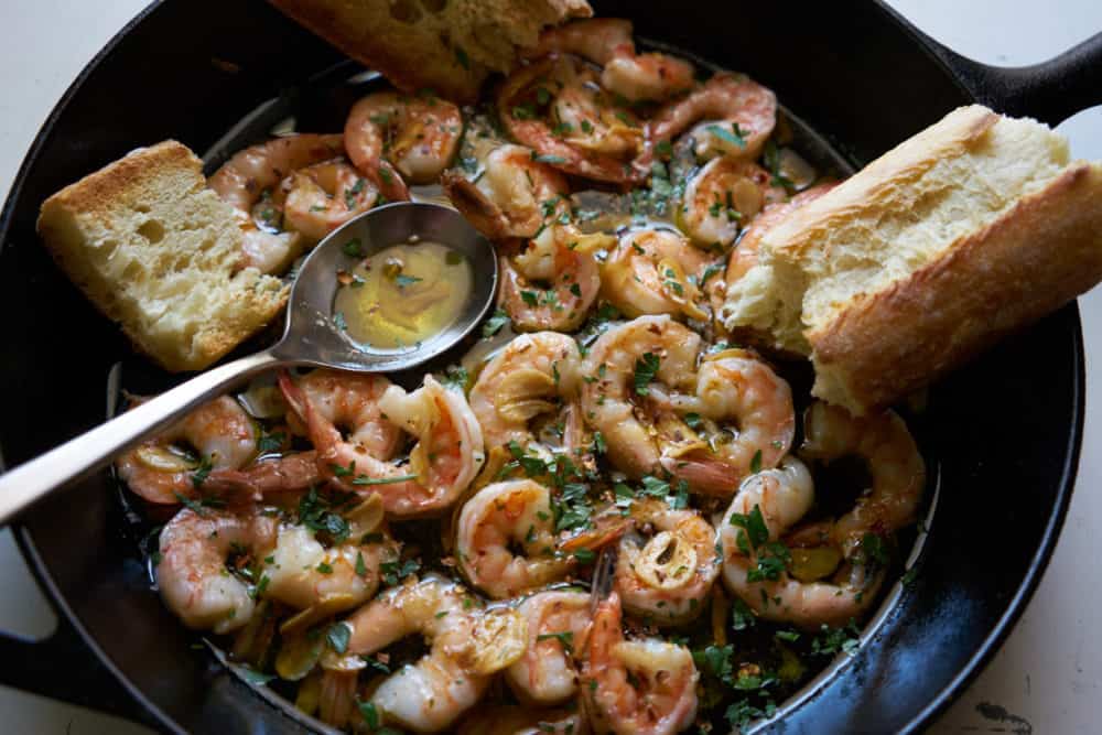 A cast iron skillet filled with garlic shrimp (gambas al ajillo). A silver spoon and torn french bread are also in the pan.