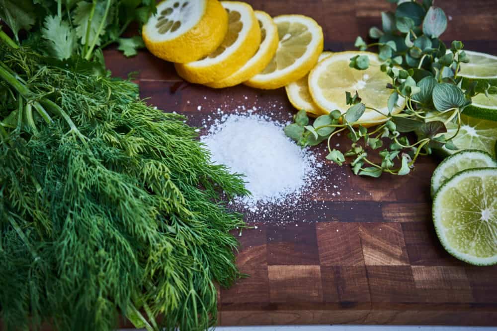 Fresh cilantro, dill and oregano on a wooden cutting board with some kosher salt and sliced lemons and limes.