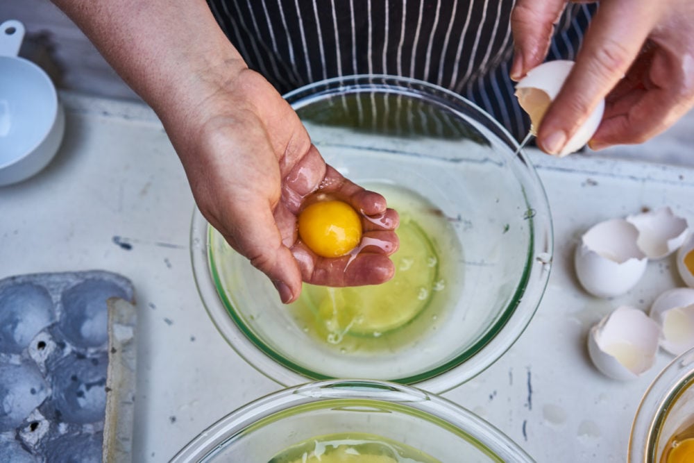 A woman's hands are shown separating and egg over a glass bowl on a white surface. 