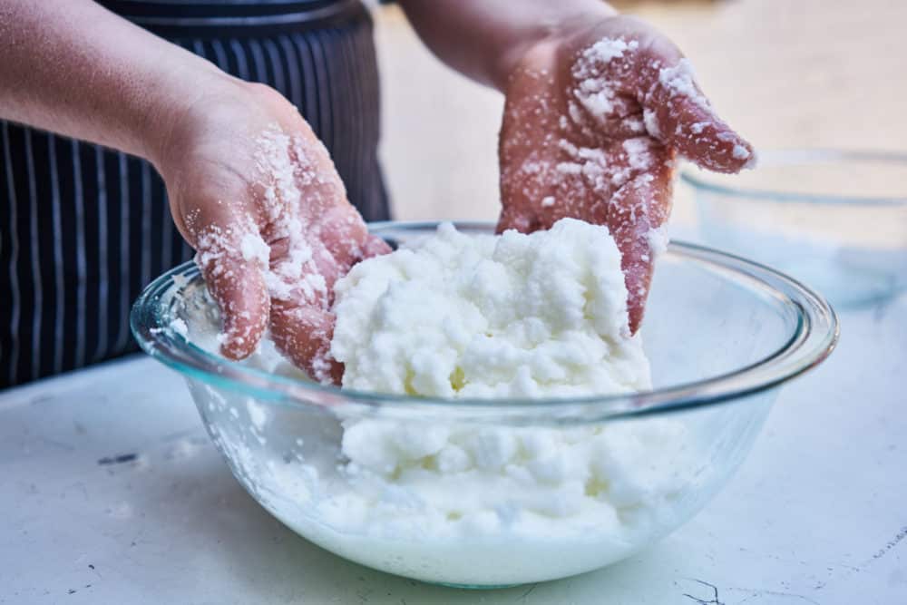 A woman's hands are shown working with a kosher salt and egg white mixture in glass bowl on a white table top. 