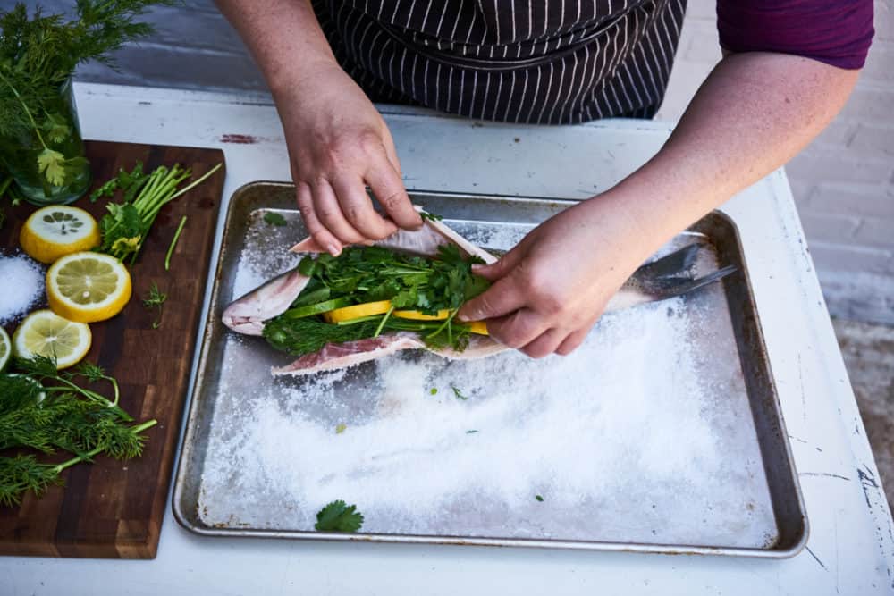 A woman stuffs a whole fish with lemon, lime and fresh herbs. The fish is laying on a bed of salt on a sheet pan. 