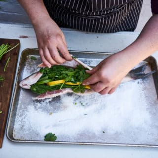 A woman's hands are shown preparing a salt baked whole fish, she stuffs a whole fish with lemon, lime and fresh herbs. The fish is laying on a bed of salt on a sheet pan.
