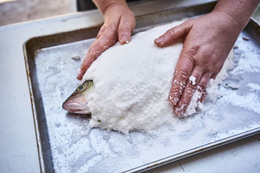 A woman's hands are shown spreading a kosher salt and egg white mixture onto a whole fish. The fish's head is exposed. 