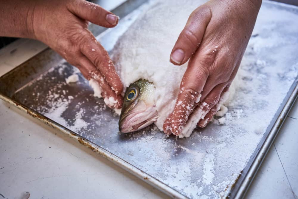 A woman's hands are shown spreading a kosher salt and egg white mixture onto a whole fish. The fish's head is exposed.