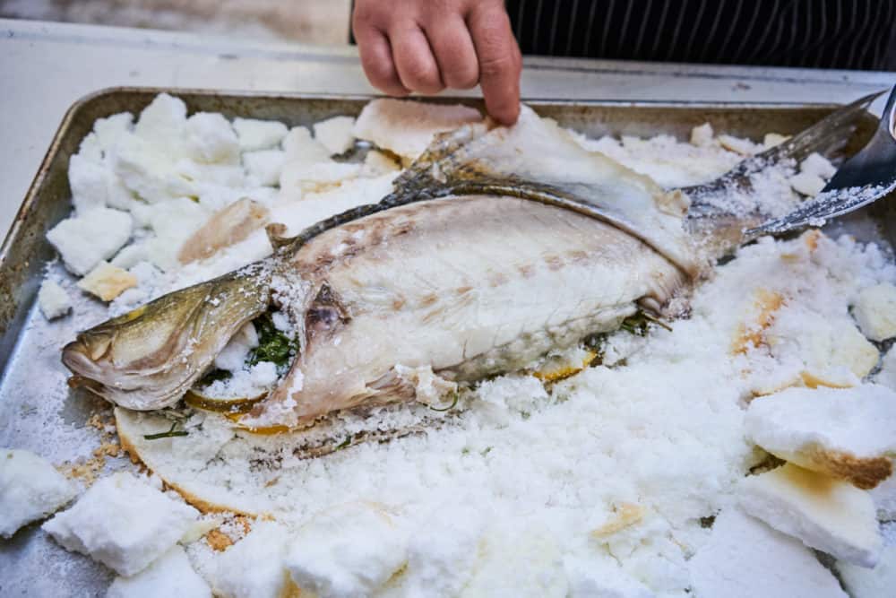 A woman's hands are shown peeling back the skin of a whole fish that has been baked in salt. 