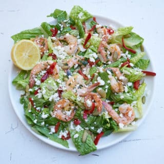 Large round plate of green salad with shrimp, romaine lettuce, cherry peppers, pepitas and feta cheese, garnished with a half a lemon.
