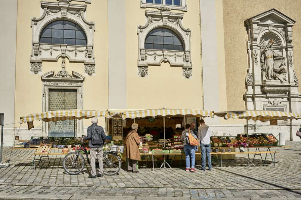 Men and women buying goods from a small produce stand with yellow and white striped awnings in Vienna. It is on a cobble-stoned street with a pale yellow Beaux Arts style building in the background.