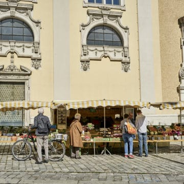 Men and women buying goods from a small produce stand with yellow and white striped awnings in Vienna. It is on a cobble-stoned street with a pale yellow Beaux Arts style building in the background.