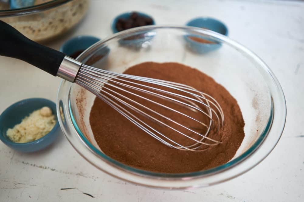 A glass bowl with a whisk, cocoa powder, almond flour and instant espresso powder. 