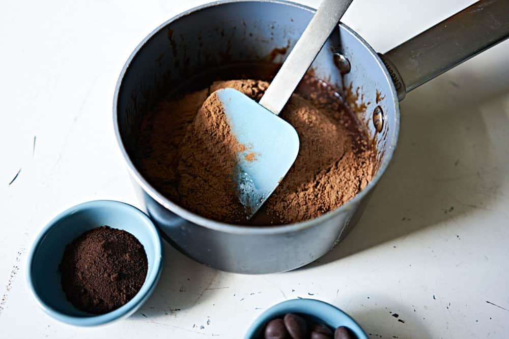 A small sauce pan holds chocolate ganache, cocoa powder, and a blue spatula. 