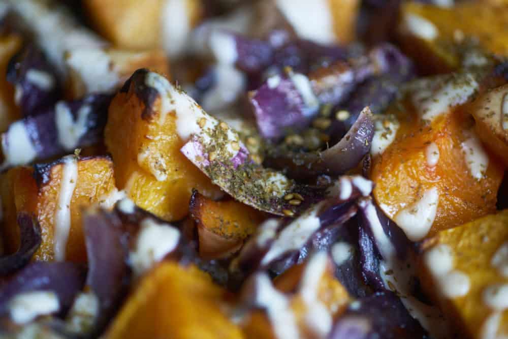 Closeup of roasted butternut squash with red onion, tahini sauce and za'atar spice based on a recipe from the cookbook Jerusalem by Ottolenghi and Tamimi displayed on a white plate.