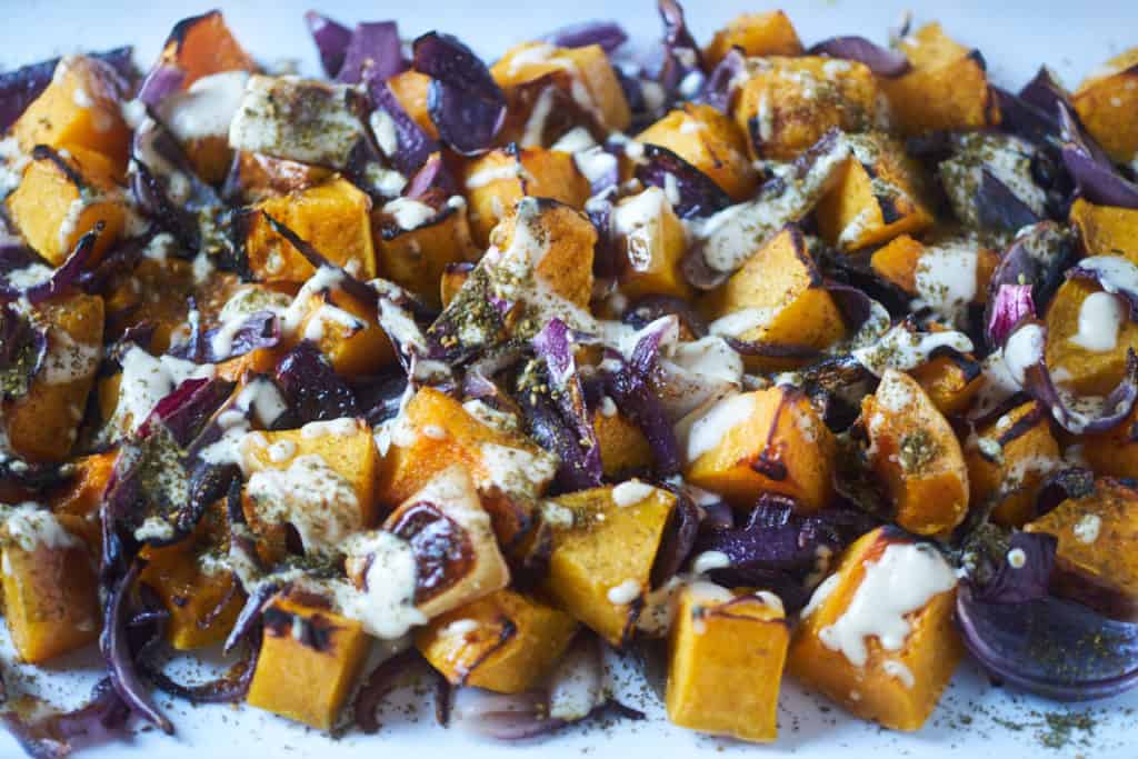 Closeup of roasted butternut squash with red onion, tahini sauce and za'atar spice based on a recipe from the cookbook Jerusalem by Ottolenghi and Tamimi displayed on a white plate.