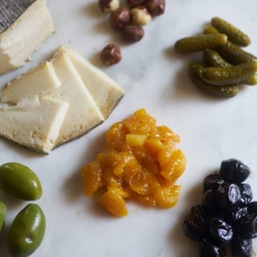 A variety of ingredients for a cheese plate including mostarda, olives, pickes, hazelnuts and cheese, on a marble surface.