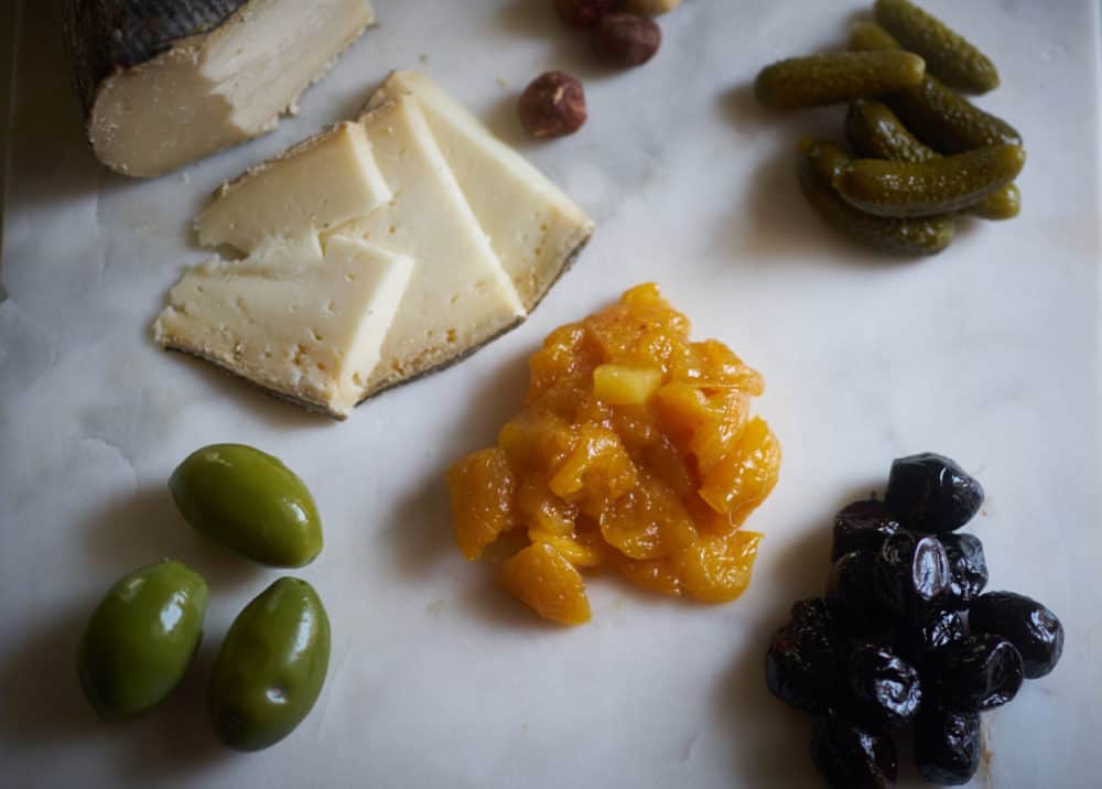 A variety of ingredients for a cheese plate including mostarda, olives, pickes, hazelnuts and cheese, on a marble surface. 