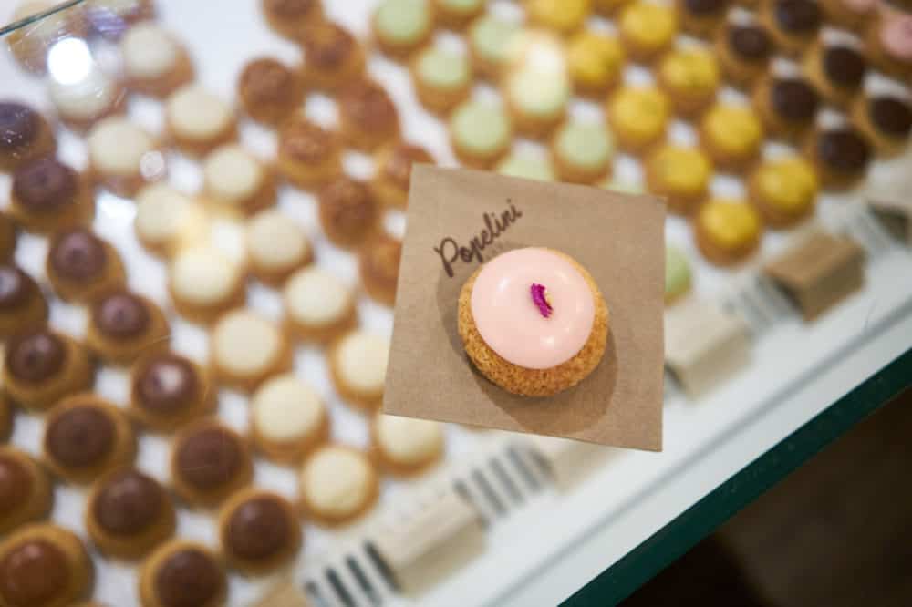 A small rose and raspberry flavored cream puff with pink icing and a dried rose petal on top is on a brown paper napkin that says Popelini. The napkin is on top of a glass display case containing more cream puffs of various colors and flavors. 