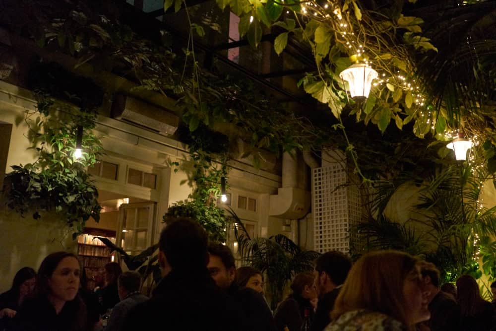 The dining room at Hotel Amour in Paris. The glass walls and ceiling are lined with plants, greenery and strings of lights. People are sitting at bistro tables throughout the dimly lit room.