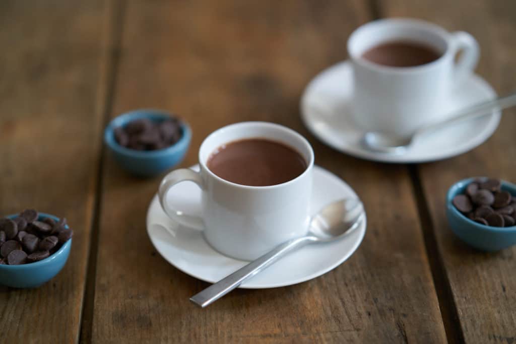 Two white cups filled with French-style hot chocolate, or chocolate chaud, surrounded by three small blue bowls filled with dark chocolate chips