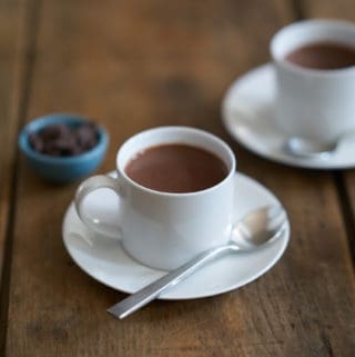Two white cups filled with French-style hot chocolate, or chocolate chaud, surrounded by three small blue bowls filled with dark chocolate chips
