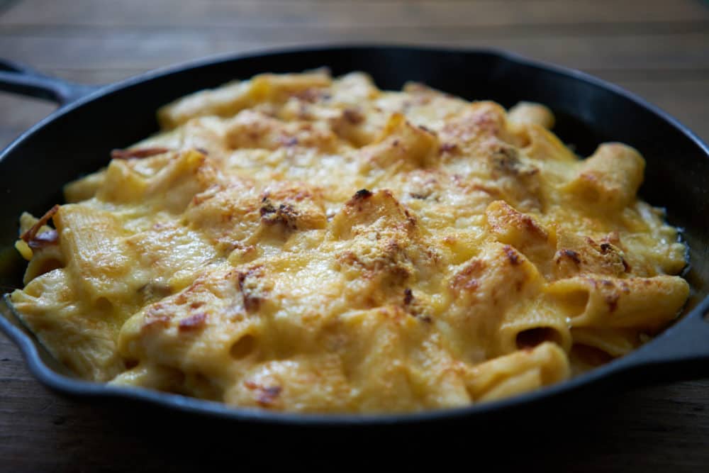 Creamy pasta with bacon and comté cheese (Pâtes aux lardons et comté) in a cast iron pan on a wooden table. The cheese on top is golden brown.