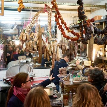 The interior of Caractère de Cochon, a charcuterie in the Marais neighborhood in Paris. Sausages and dried peppers are hanging from the ceiling. A tour guide and tour group is in front of the counter as a man slices charcuterie behind it.
