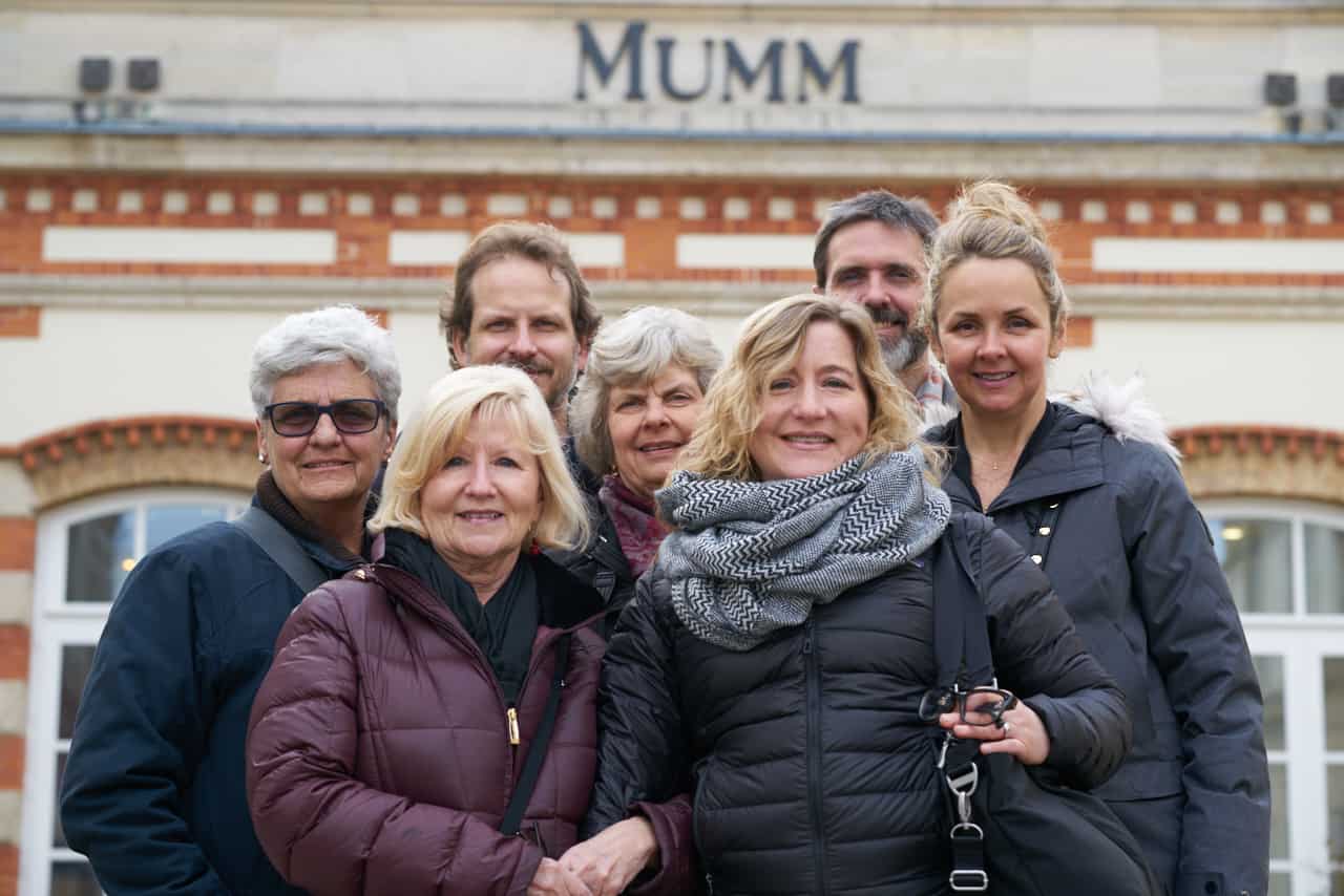 A family of seven adults wearing winter coats are smiling for a portrait in front of G.H. Mumm in Reims, France. There are two men and five women.