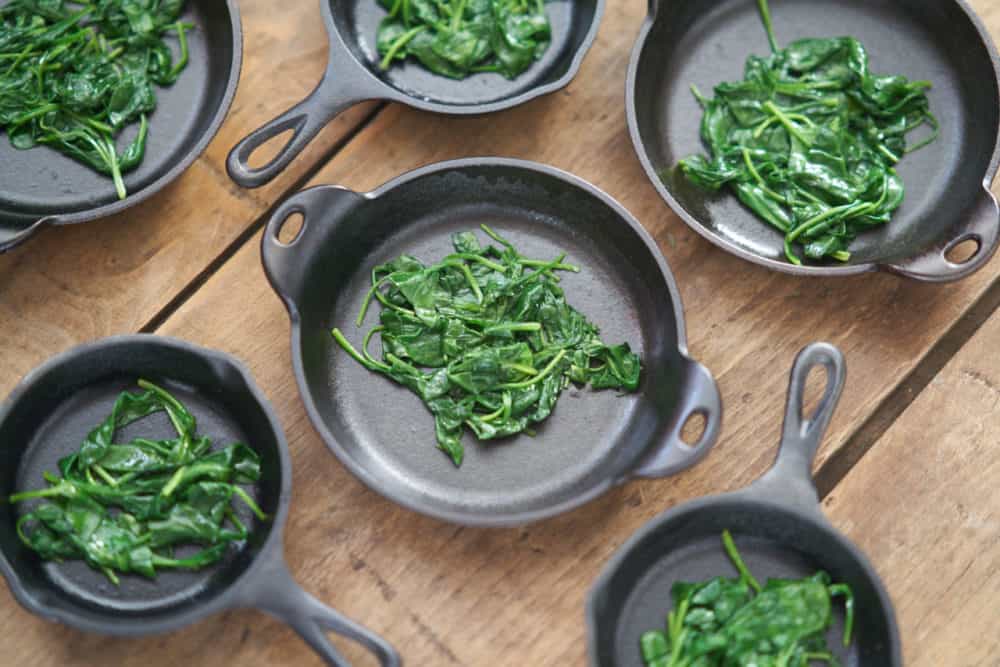 Several small cast-iron skillets and serving dishes containing a small amount of cooked spinach are displayed on a wooden surface. 