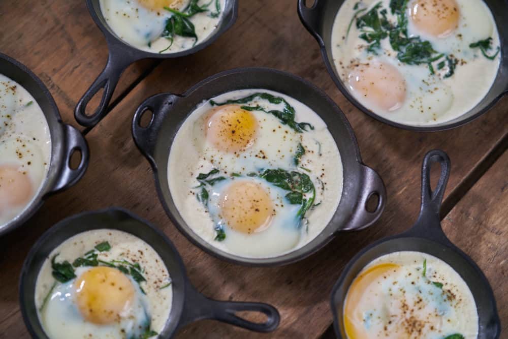 Several servings of eggs en cocotte with spinach in small cast-iron skillets and serving dishes are waiting to be topped with bacon. They are displayed on a wooden surface. 
