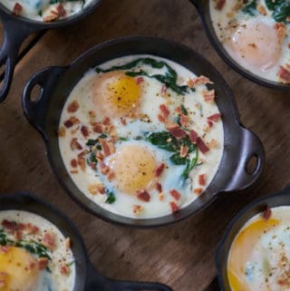 Several servings of eggs en cocotte with spinach and bacon in small cast-iron skillets and serving dishes are displayed on a wooden surface.