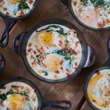 Several servings of eggs en cocotte with spinach and bacon in small cast-iron skillets and serving dishes are displayed on a wooden surface.