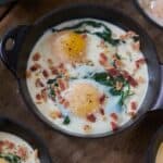 Eggs en cocotte with spinach and bacon in small cast-iron skillets and serving dishes, displayed on a wooden surface.