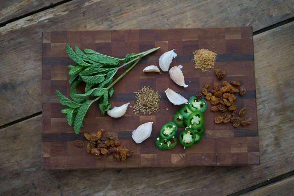Sliced jalapeños, golden raisins, garlic cloves, fresh mint, cumin seeds, and ground coriander on a wooden cutting board.