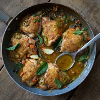 A skillet with chicken escabeche sits on a wooden surface. A sliver spoon is in the pan on the right side. Garlic, golden raisins and jalapeños can be seen in the escabeche sauce, and the dish is garnished with fresh mint.
